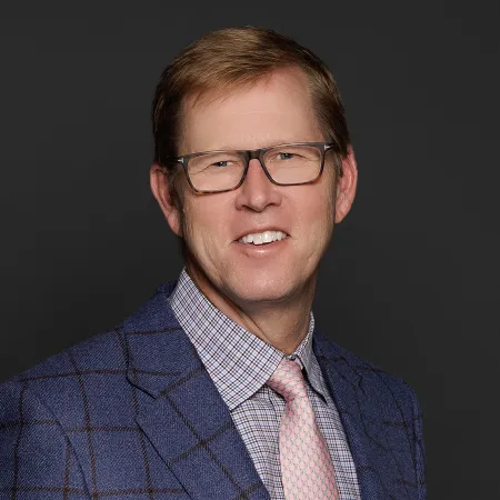 Middle-aged man with glasses wearing a blue checked blazer, plaid shirt, and pink tie smiling against black background