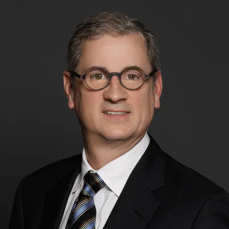 Middle-aged man wearing glasses, a black suit, white shirt, and striped tie against a dark background