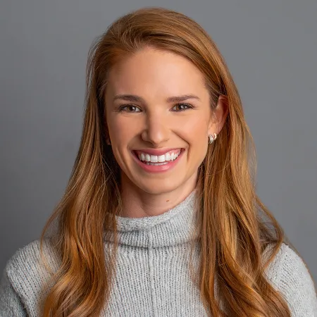 Smiling woman with long red hair wearing a gray knit sweater against a plain gray background