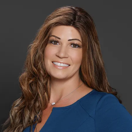 Smiling woman with long wavy brown hair wearing a blue top and a delicate necklace on gray background