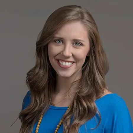 Smiling woman with long brown hair wearing a blue top and yellow beaded necklace on gray background