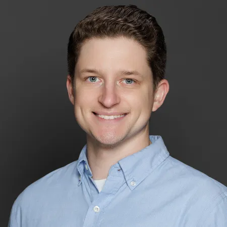 Young man with curly brown hair and blue eyes smiling wearing a light blue shirt on gray background