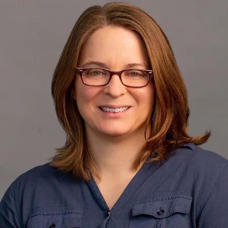 Smiling woman with shoulder-length brown hair wearing glasses and a blue shirt against a gray background