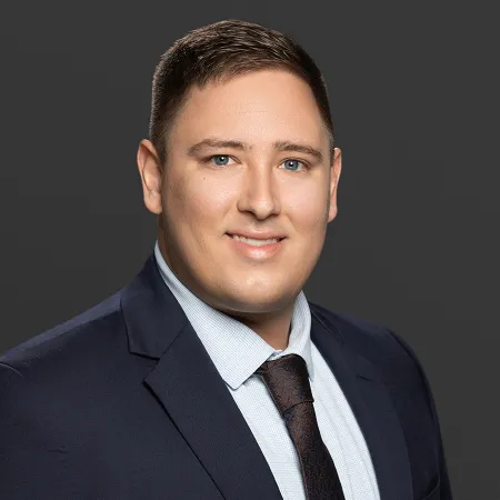 Professional headshot of a smiling businessman in a navy suit and patterned tie against a dark gray background