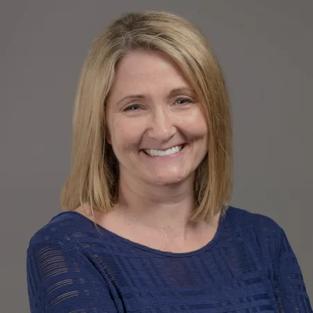Smiling middle-aged woman with blonde hair wearing a textured navy blue top against a gray background