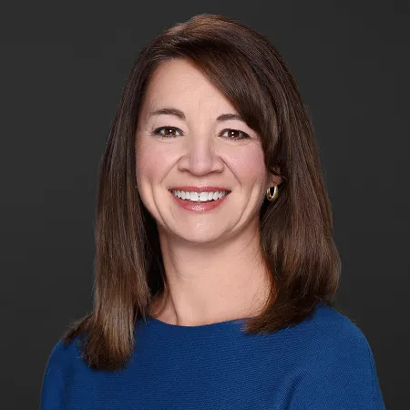 Smiling woman with straight brown hair wearing blue top and gold hoop earrings on a dark background