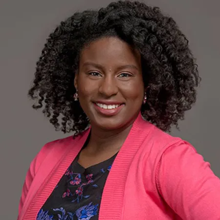Smiling woman with natural curly hair wearing a pink cardigan over a floral top against a plain background