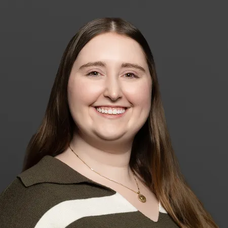 Smiling young woman with long brown hair wearing a striped sweater and gold necklace against a gray background