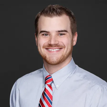 Smiling young man with short hair and beard wearing light blue shirt and red striped tie on dark background
