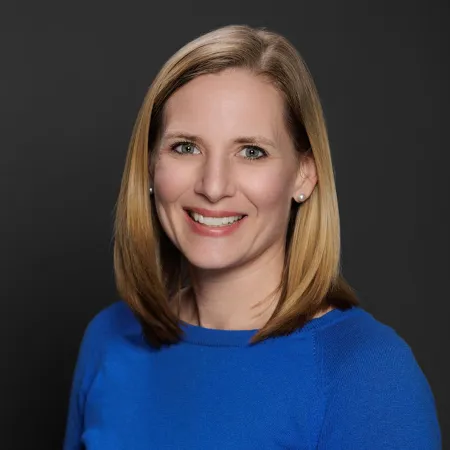 Smiling woman with shoulder-length blonde hair wearing a blue sweater against a dark background