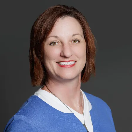 Smiling woman with short brown hair wearing a blue sweater and white shirt against a gray background