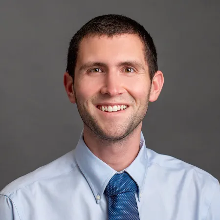 Smiling man with short dark hair wearing a light blue shirt and blue patterned tie on gray background