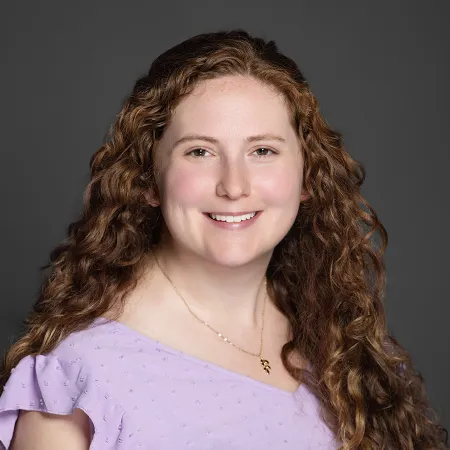 Smiling woman with long curly red hair wearing a light purple top and gold necklace against a gray background