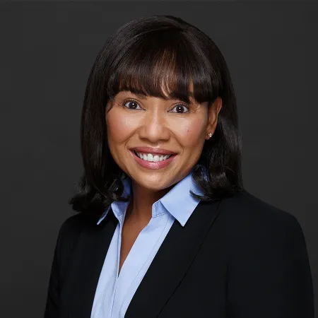 Smiling professional woman with dark hair wearing a black blazer and light blue collared shirt on gray background