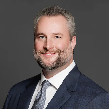 Smiling man with gray hair and beard wearing a navy suit and patterned tie against a gray background