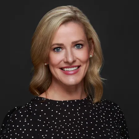 Smiling blonde woman with blue eyes wearing pearl earrings and a black polka dot blouse on dark background