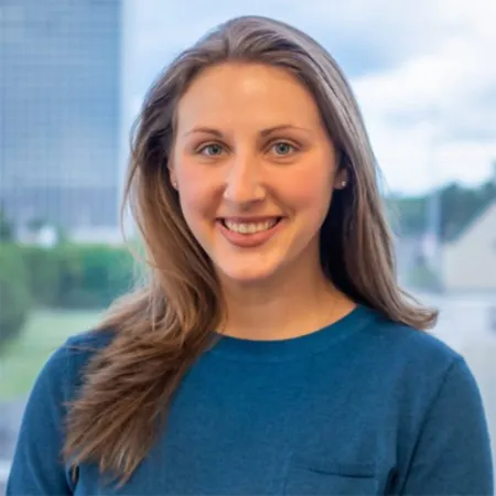 Smiling young woman with long light brown hair wearing a blue sweater standing outdoors with buildings in the background