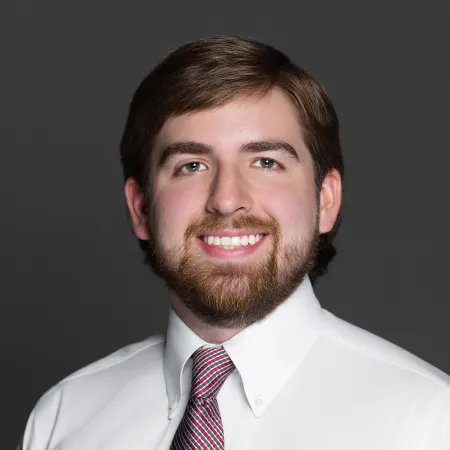 Smiling man with brown hair and beard wearing white shirt and red striped tie against gray background