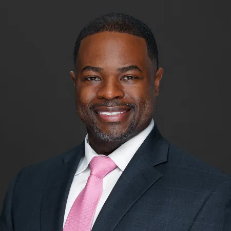 Smiling man in a dark suit and pink tie against a plain dark background headshot