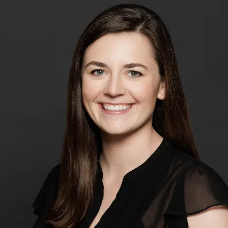 Smiling young woman with long brown hair wearing a black top against a dark gray background.