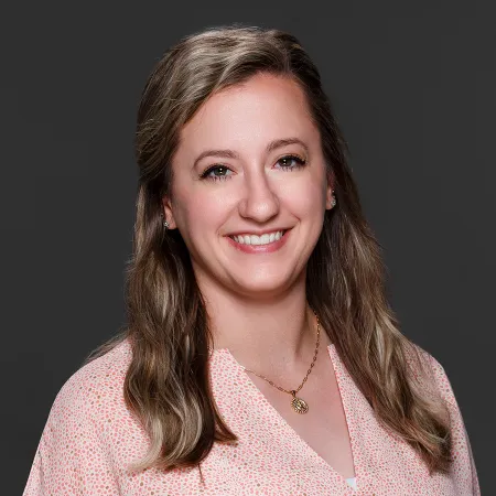 Smiling woman with wavy light brown hair wearing a pink patterned blouse and gold necklace on gray background