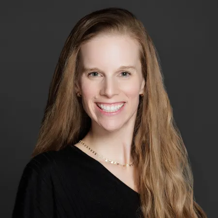 Smiling woman with long blonde hair wearing a black top and gold necklace on a dark gray background.