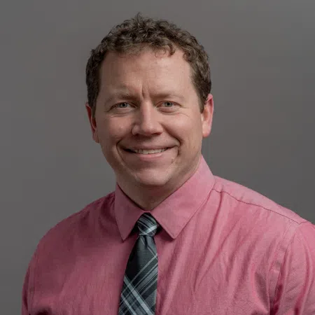 Smiling man with curly hair wearing a pink shirt and plaid tie posing against a gray background