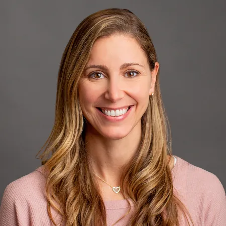 Smiling woman with long blonde hair wearing a pink top and heart-shaped necklace against a gray background