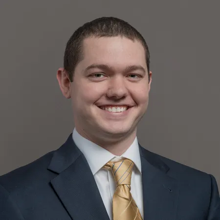Young man smiling in a navy suit with a white shirt and gold striped tie against a gray background