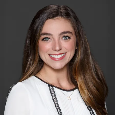 Smiling woman with long brown hair wearing white blouse with black trim and gold necklace on gray background