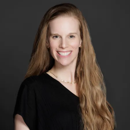 Smiling woman with long blonde hair wearing a black top and delicate necklace on dark gray background