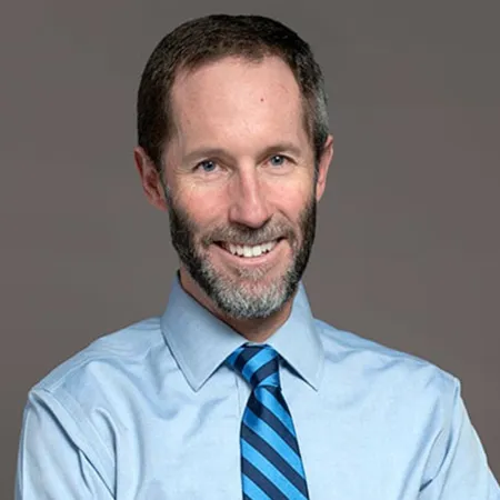 Smiling man with beard wearing light blue shirt and blue striped tie against gray background