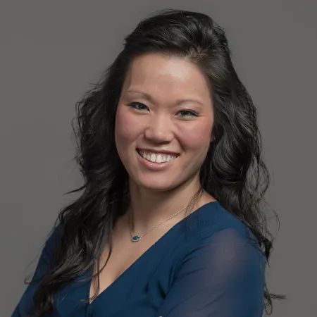 Smiling woman with long black hair wearing blue blouse and pendant necklace against plain gray background
