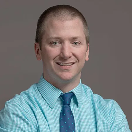 Smiling man in turquoise checkered shirt and navy blue tie with polka dots against a plain gray background