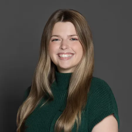 Smiling woman with long blonde hair wearing a dark green short-sleeve sweater against a gray background
