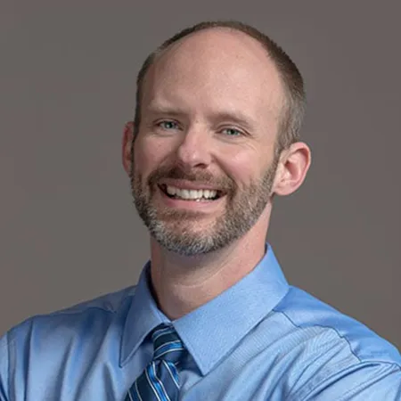 Smiling man with a beard wearing a blue dress shirt and striped tie against a plain gray background