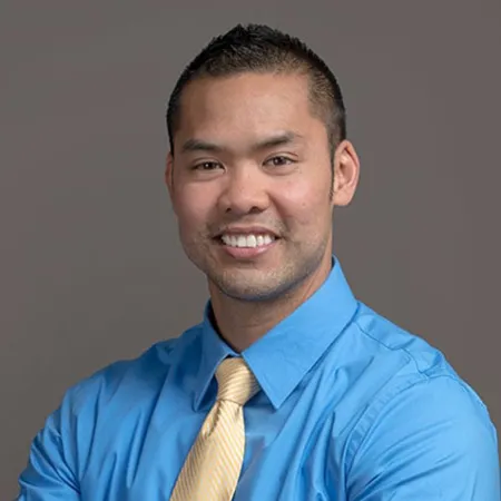 Smiling man in blue shirt and yellow tie posing with folded arms against a gray background