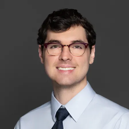 Smiling young man in glasses wearing a light blue shirt and dark tie against a gray background