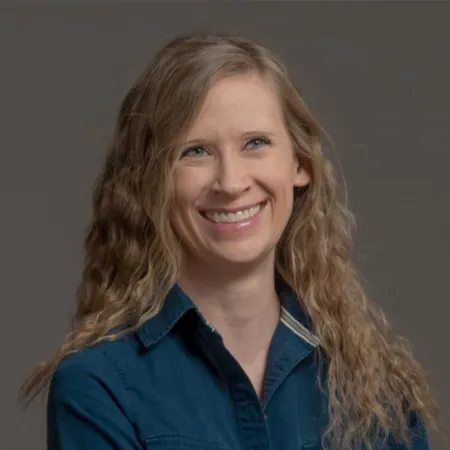 Smiling woman with long curly blonde hair wearing a blue collared shirt against a gray background