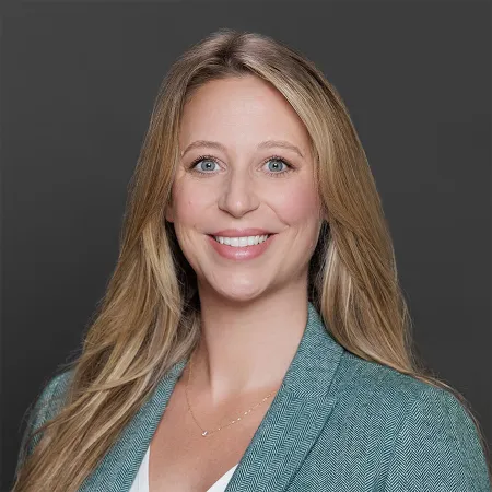 Smiling blonde woman with blue eyes wearing a teal blazer and white top against a plain dark background