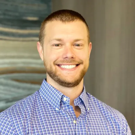 Smiling man with short hair and beard wearing a blue checkered shirt against a blurred background.
