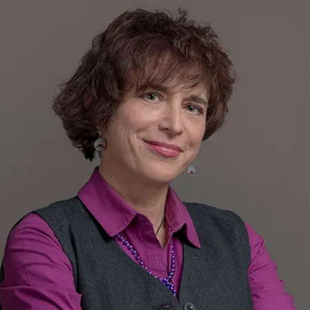Smiling woman with short curly hair wearing a purple shirt, dark vest, and purple necklace against gray background