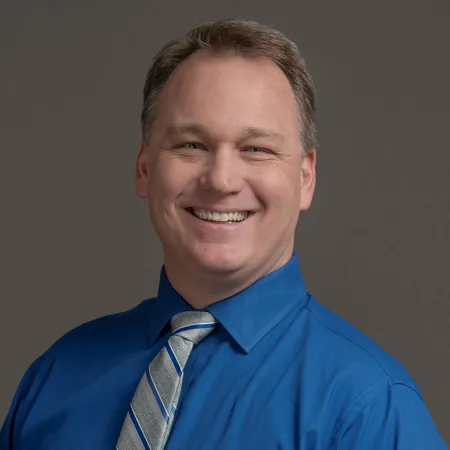 Smiling man wearing a blue shirt and striped tie against a neutral gray background