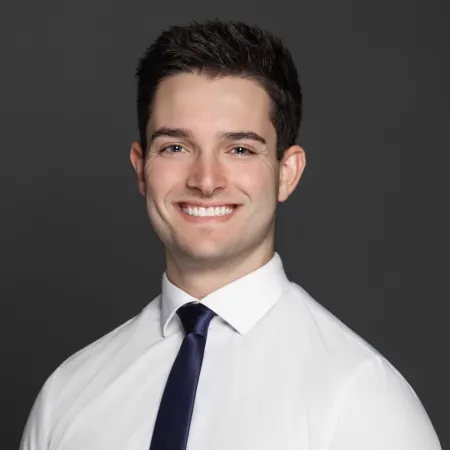 Young man in white shirt and dark tie smiling against a dark gray background, professional headshot.