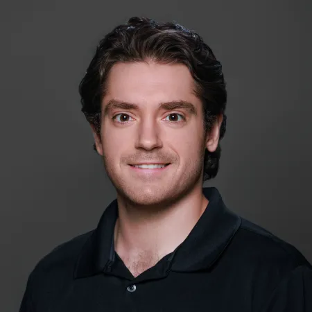 Smiling young man with dark curly hair wearing a black polo shirt against a gray background