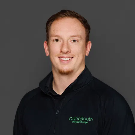 Smiling man wearing black OrthoSouth Physical Therapy jacket against a gray background