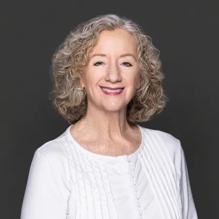 Smiling middle-aged woman with curly hair wearing a white blouse against a dark gray background