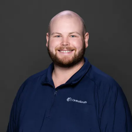 Smiling bald man with beard wearing a navy blue OrthoSouth quarter-zip jacket against gray background