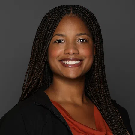 Smiling woman with long braided hair wearing an orange top and black jacket against a dark gray background.
