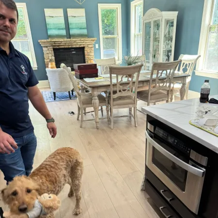 Curly-haired dog holding a toy in a bright kitchen and dining room with wood flooring and blue walls.